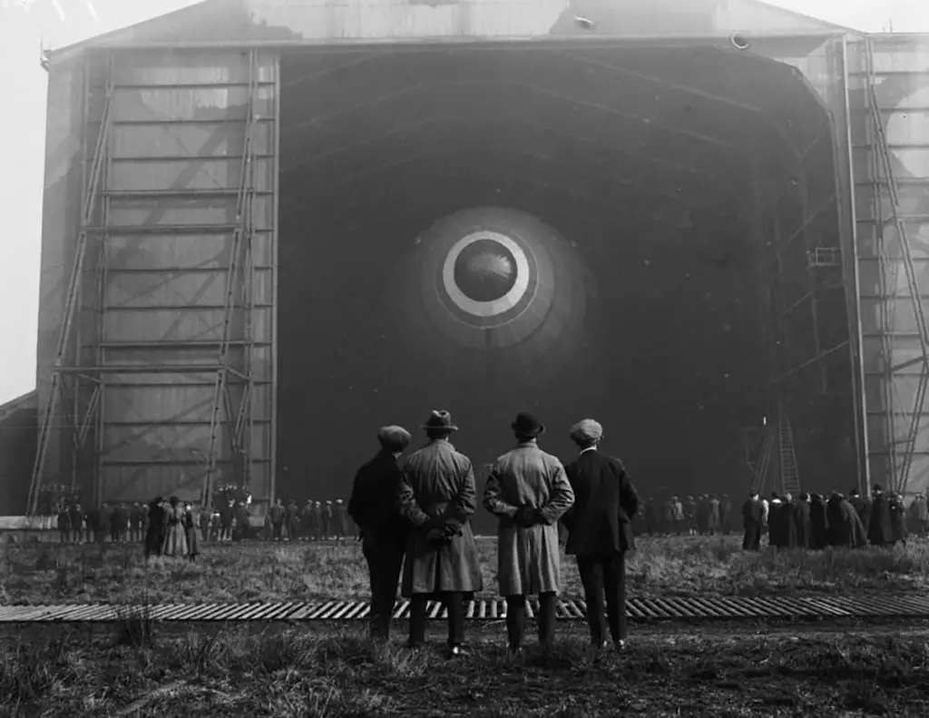 Fotografía de un grupo de personas viendo a un dirigible salir de un hangar.